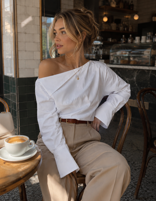 Woman wearing a white one-shoulder long sleeve blouse with beige trousers at a café.